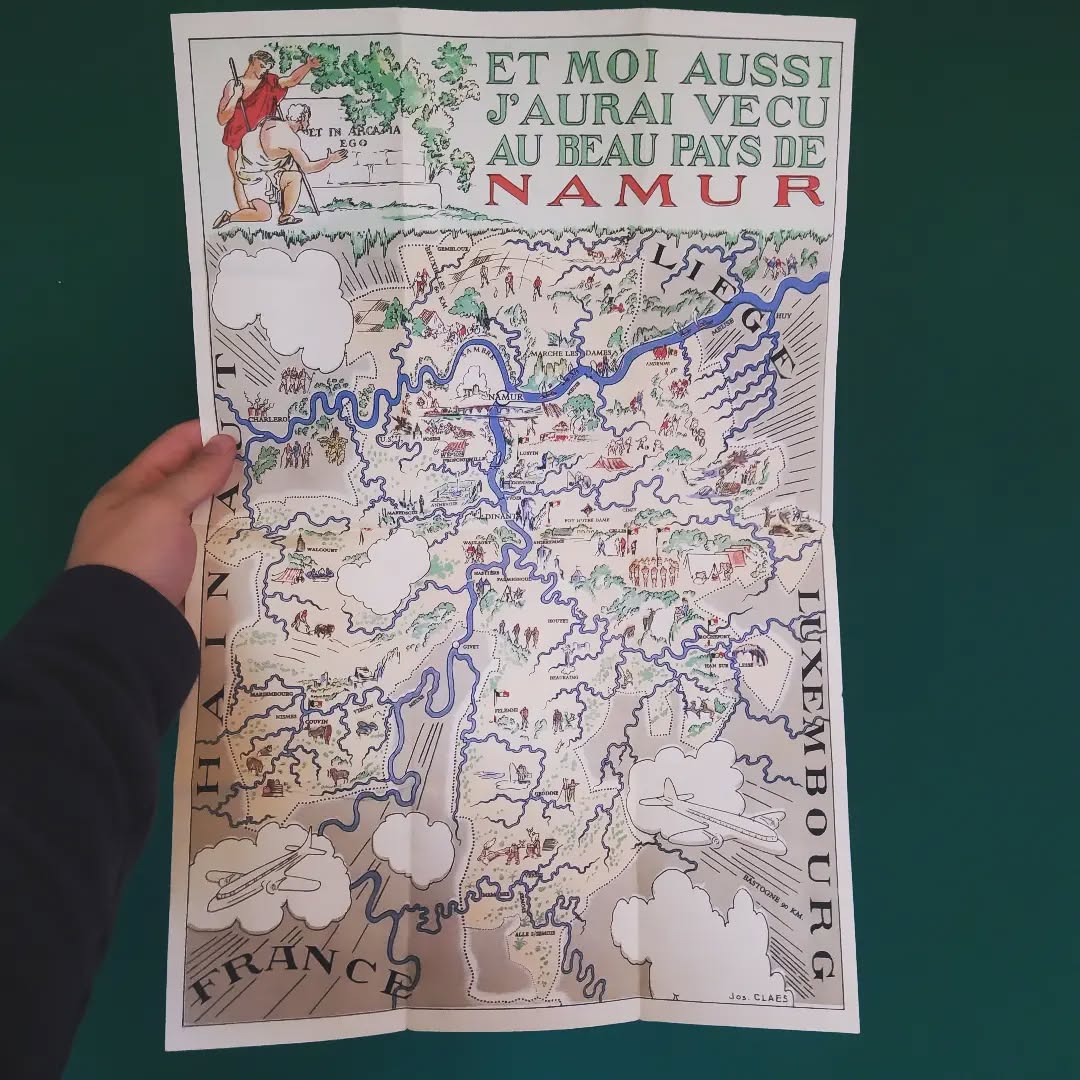 Affiche géographique touristique et illustrée complète de la province de Namur en Belgique. L'affiche, intitulée "ET MOI AUSSI J'AURAI VECU AU BEAU PAYS DE NAMUR", montre une carte dessinée avec les rivières Meuse et Sambre, ainsi que de nombreuses villes et illustrations de scènes de vie et d'activités. L'auteur est Joe Claes et elle date des années 1950. Elle est visible sur toute sa hauteur, posée sur un fond vert foncé, tenue par une main.