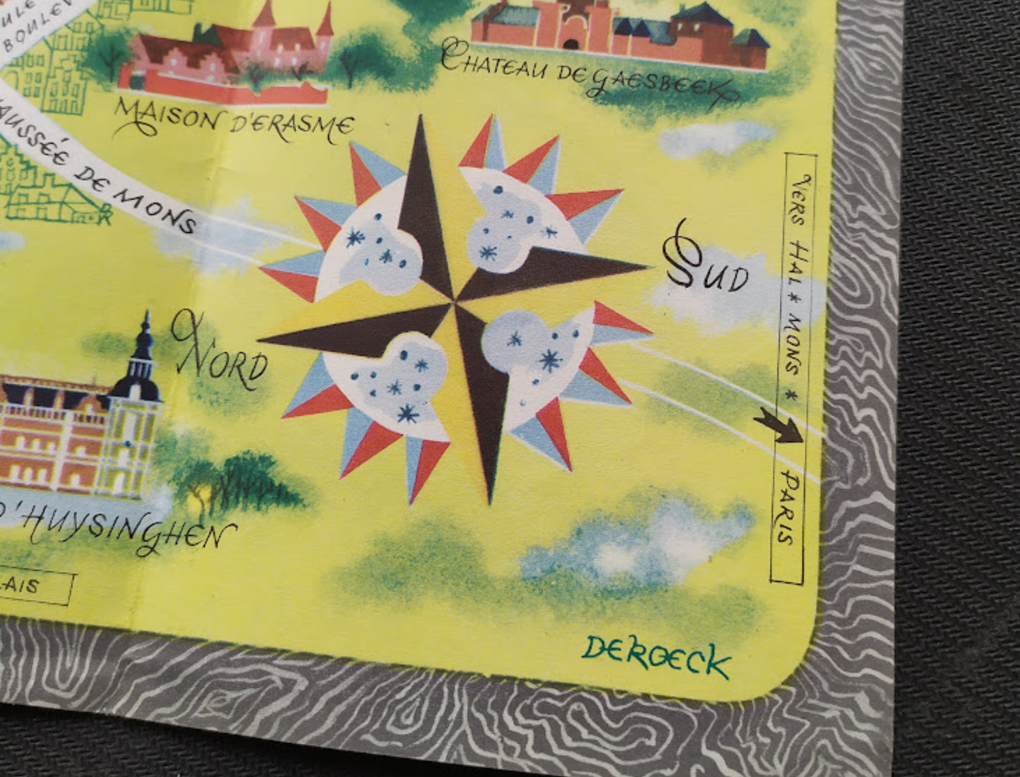 Détail du coin inférieur droit présentant une rose des vents géométrique colorée et la signature de l'artiste "De Roeck" sur un fond jaune avec des nuages stylisés.